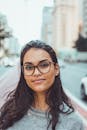 A confident woman wearing glasses smiles subtly while standing on an urban street with blurred background.